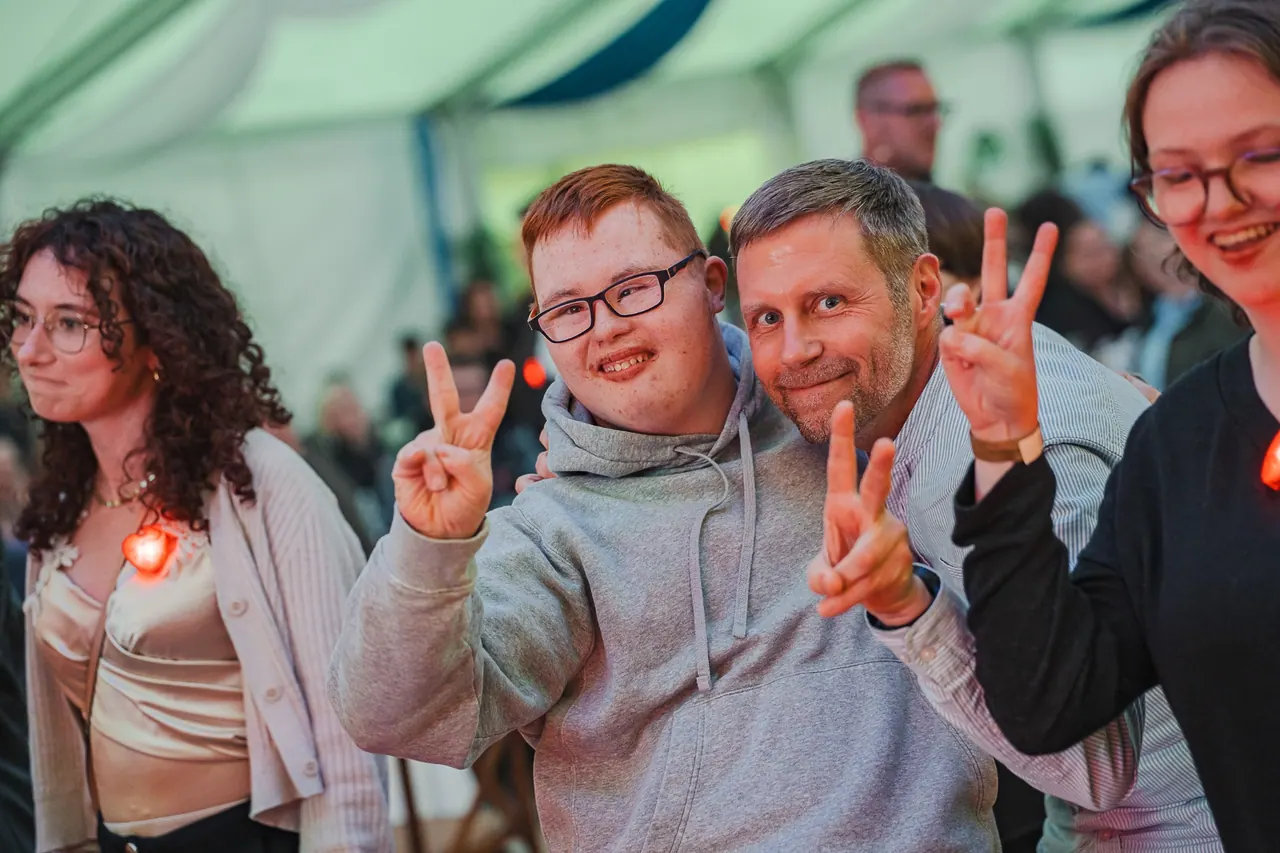 R+V Herzmomente 2025 — Junger Mann mit Down-Syndrom und Freund zeigen Peace-Zeichen auf der Tanzfläche