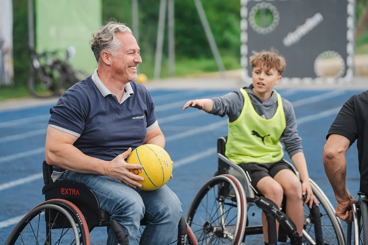 R+V Herzmomente 2025 — Mann im Sportrollstuhl mit Basketball im Gespräch mit Jugendlichem